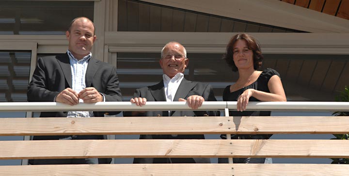 Équipe professionnelle souriante sur un balcon moderne Trois professionnels souriants, deux hommes en costume et une femme, posant sur un balcon derrière une balustrade.