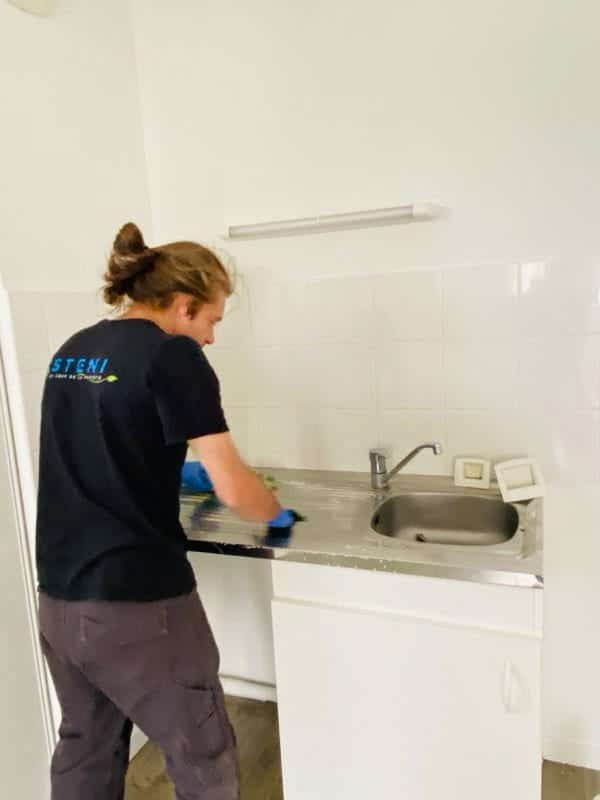Agent d'entretien STENI nettoie une cuisine Professionnel STENI nettoie un plan de travail en inox dans une cuisine blanche avec gants bleus.